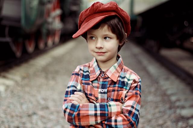 kilkuletni chłopiec o ciemnych włosach ubrany w koszulę w kratkę i czerwony kaszkiet stoi z założonymi rękami i patrzy w bok