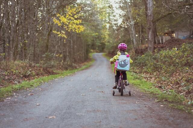 dziewczynka z kolorowym plecakiem w żółtej kurtce i z różowym kaskiem  jadąca tyłem leśną ścieżką na rowerku z czterema kołami