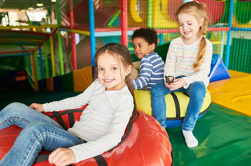Lokalizacja dziecka w Play, czyli troje dzieci, dwie dziewczynki w jeansach i jasnych bluzkach oraz chłopiec w bluzce w paski i ciemnych włosach, bawią się w bawialni na miękkich, kolorowych matach