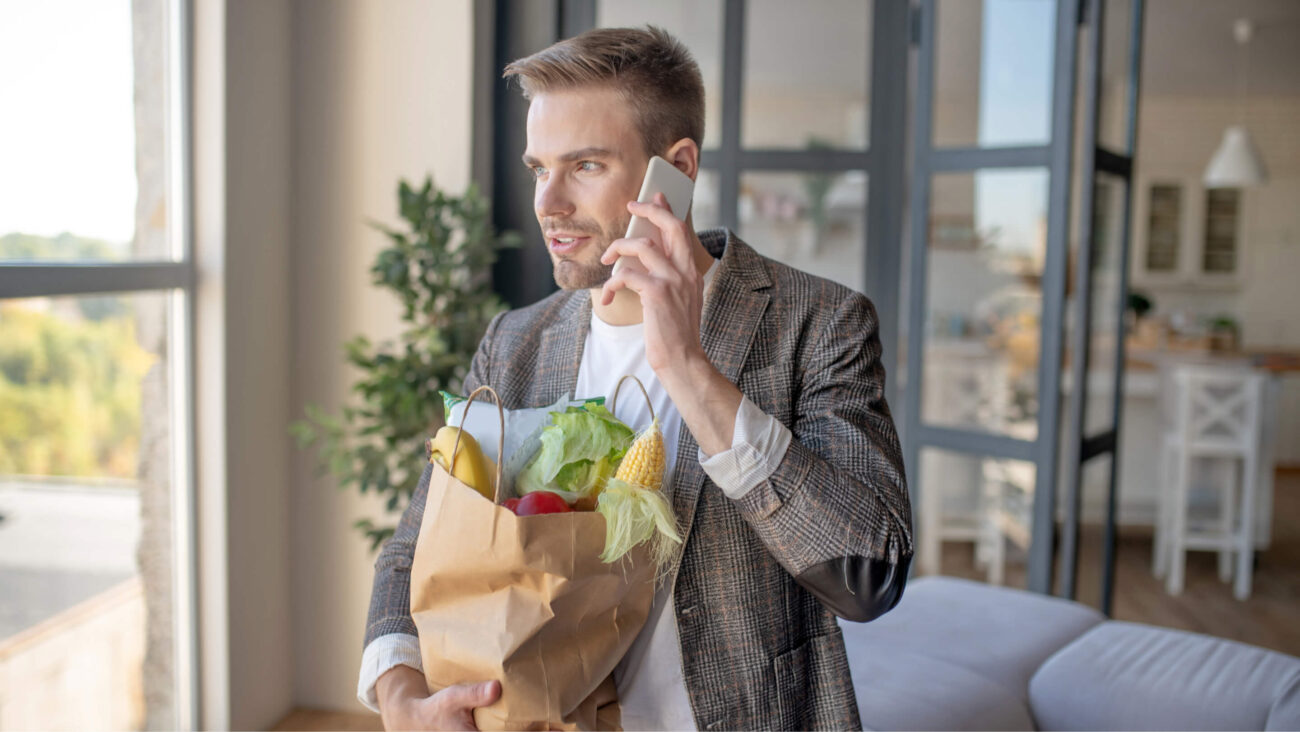 Mężczyzna korzystający z aplikacji do zamawiania jedzenia przez internet, trzymający w rękach zakupy w papierowej torbie
